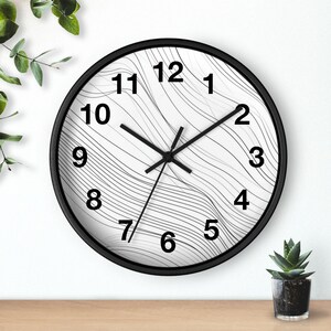 May include: A round wall clock with a black frame and hands. The clock face is white with black numbers and a wavy, striped pattern. The clock is on a wooden surface with plants in the background.