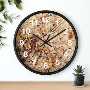 Puede incluir: Reloj de pared redondo con marco negro y esfera beige con diseño abstracto colorido. La esfera del reloj tiene números blancos y manecillas negras. El reloj mide aproximadamente 30 cm de diámetro.