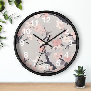 May include: Round wall clock with a black frame. The clock face features a gray background with a floral pattern of pink and white blossoms and dark gray branches. White numerals mark the hours, and black hands indicate the time.
