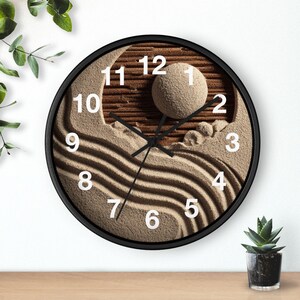 May include: A circular wall clock with a black frame. The clock face has a sand design with a sphere and wavy lines. White numerals mark the hours. The clock hands are black.