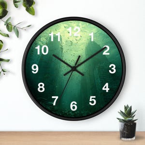 Puede incluir: Un reloj de pared redondo con marco negro. La esfera del reloj presenta una escena submarina con edificios y peces. Los números blancos marcan las horas, y las manecillas negras indican la hora.