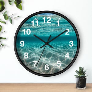 May include: A round wall clock with a black frame. The clock face features an underwater scene with blue water and white sand. White numerals mark the hours, and black hands indicate the time. The clock is a decorative item.