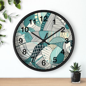 Könnte beinhalten: Eine runde Wanduhr mit schwarzem Rahmen. Das Zifferblatt hat ein abstraktes Design in Türkis, Weiß und Schwarz mit den Zahlen 1 bis 12. Die Zeiger der Uhr sind schwarz.