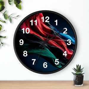 Puede incluir: Un reloj de pared redondo con marco negro. La esfera del reloj presenta un diseño abstracto colorido con tonos rojos, azules y verdes. Los números blancos marcan las horas. Las manecillas del reloj son negras.