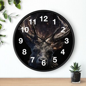 May include: A round black wall clock featuring a detailed image of a stag's head with large antlers against a dark background. The clock face has white numerals and hands, and the clock is surrounded by a black frame.