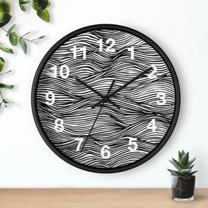 Puede incluir: Un reloj de pared redondo con marco negro. La esfera del reloj presenta un patrón ondulado en blanco y negro con números blancos. Las manecillas del reloj son negras. El reloj está montado en una pared blanca.