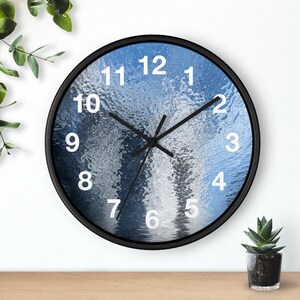 Op de afbeelding: Ronde wandklok met een zwart frame. De wijzerplaat heeft een blauw-wit waterachtig ontwerp met witte cijfers. Zwarte wijzers geven de tijd aan. Op de achtergrond is een kleine vetplant in een pot te zien.