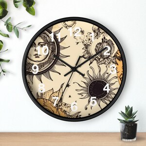 May include: A round wall clock with a black frame. The clock face features a cream-colored background with sun and floral illustrations in black and white. White numerals mark the hours. The clock hands are black.