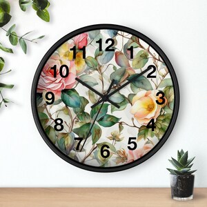 May include: Round wall clock with a black frame. The clock face features a floral design with pink and yellow roses, green leaves, and a white background. Black hands indicate the time. The clock is on a wooden surface.
