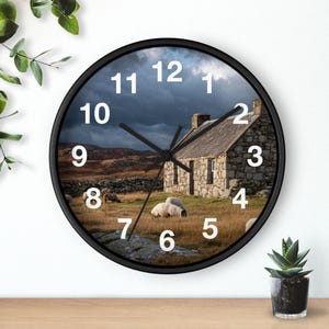 May include: A round wall clock with a black frame. The clock face features a scenic image of a stone cottage, sheep, and a cloudy sky. White numbers mark the hours. The clock hands are black.