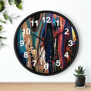 Puede incluir: Un reloj de pared redondo con marco negro. La esfera del reloj presenta un diseño abstracto colorido con vetas azules, rojas, marrones y blancas. Los números blancos marcan las horas. El reloj está sobre una superficie de madera.