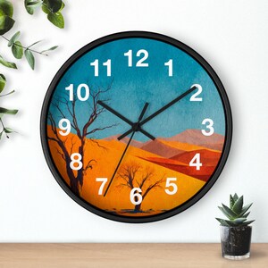 Op de afbeelding: Ronde wandklok met een zwart frame. De wijzerplaat toont een woestijnlandschap met een blauwe lucht, oranje zandduinen en kale bomen. Witte cijfers markeren de uren en zwarte wijzers geven de tijd aan.