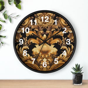 May include: Round black wall clock with a gold and black ornate design. The clock face features Roman numerals in white and black clock hands. The clock is on a wooden surface with a small potted plant.