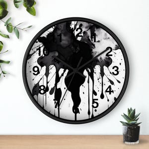 Puede incluir: Un reloj de pared redondo con marco negro y esfera blanca. La esfera del reloj presenta un diseño de salpicaduras de tinta negra con efecto de goteo de pintura. Los números negros marcan las horas. Las manecillas del reloj son negras.