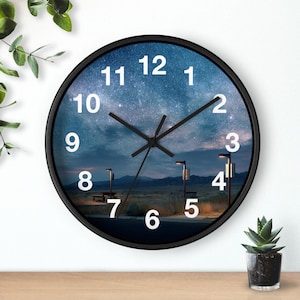 May include: Round black wall clock featuring a starry night sky design. The clock face displays white numerals and black hands. The background image shows a landscape with streetlights and a bench.