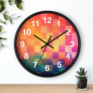May include: Round wall clock with a black frame. The clock face features a colorful, checkered pattern in shades of orange, yellow, pink, and green. White numerals mark the hours, and black hands indicate the time.