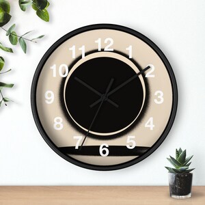 May include: A round wall clock with a black frame. The clock face is beige with a black circle in the center and white numerals. Black hands indicate the time. A black horizontal stripe is at the bottom.