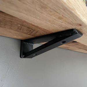 Pode incluir: Suporte de prateleira de metal preto fixado a uma prateleira de madeira. O suporte é angular e possui um suporte triangular. A prateleira é feita de madeira clara com grão visível.