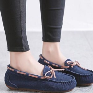 May include: Navy blue moccasin-style shoes with a suede-like texture. The shoes feature a brown leather trim and a decorative bow on the vamp. The soles are a dark brown color. These casual shoes are suitable for everyday wear.