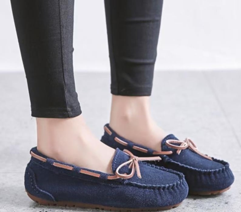 May include: Navy blue moccasin-style shoes with a suede-like texture. The shoes feature a brown leather trim and a decorative bow on the vamp. The soles are a dark brown color. These casual shoes are suitable for everyday wear.