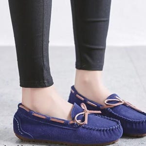 May include: A pair of navy blue suede moccasin-style loafers. The shoes have a rounded toe, a stitched vamp, and a light brown leather trim. A light brown bow is tied on the top of each shoe. The shoes are worn with black pants.