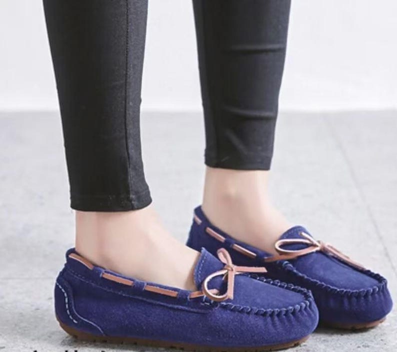 May include: A pair of navy blue suede moccasin-style loafers. The shoes have a rounded toe, a stitched vamp, and a light brown leather trim. A light brown bow is tied on the top of each shoe. The shoes are worn with black pants.