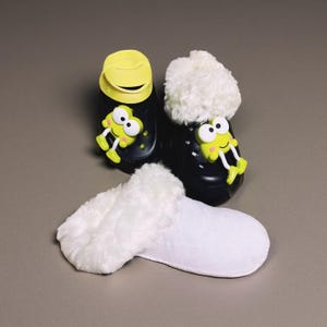 May include: Black and yellow children's clogs with a frog design and white faux fur trim. A pair of white, fluffy slippers are in front of the clogs. The clogs have a yellow strap and a frog face on the front.