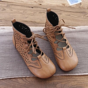 May include: A pair of tan leather ankle boots with intricate cut-out designs. The boots feature a lace-up closure and a rounded toe. The interior is black, and the boots are set on a neutral-colored surface. The overall style is bohemian.