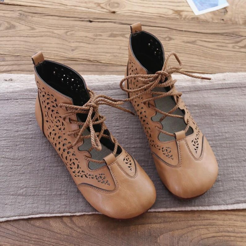 May include: A pair of tan leather ankle boots with intricate cut-out designs. The boots feature a lace-up closure and a rounded toe. The interior is black, and the boots are set on a neutral-colored surface. The overall style is bohemian.