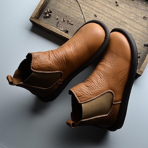 May include: A pair of brown leather Chelsea boots with elastic side panels and dark soles. The boots have a textured appearance and a pull tab at the back. The footwear is displayed on a light gray surface with a wooden tray in the background.