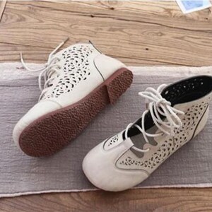 May include: A pair of cream-colored, open-toe ankle boots with intricate cut-out designs. The boots have brown soles and laces. The shoes are placed on a gray fabric and wooden surface.