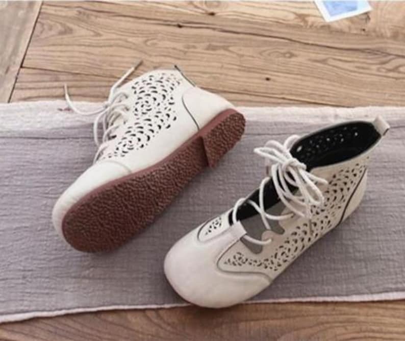 May include: A pair of cream-colored, open-toe ankle boots with intricate cut-out designs. The boots have brown soles and laces. The shoes are placed on a gray fabric and wooden surface.