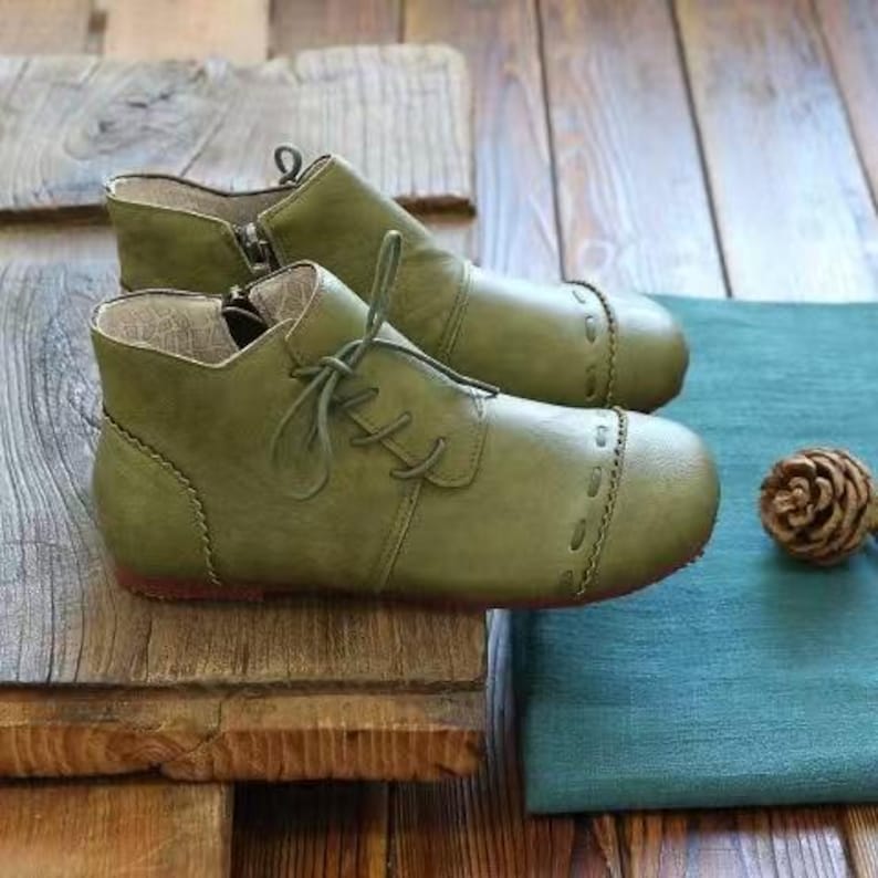 May include: A pair of olive green ankle boots with laces and a side zipper. The boots have a rounded toe and are made of a soft material. The boots are displayed on a wooden surface.