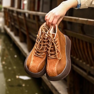 May include: A pair of brown leather ankle boots with light brown laces and a thick, dark brown sole. The boots have a soft, light-colored interior and are held up by a hand.