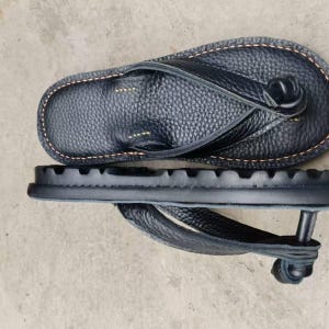 Puede incluir: Chanclas de cuero negro con superficie texturizada y costuras marr&oacute;n claro. La suela es gruesa y estriada. La correa de la chancla tambi&eacute;n es de cuero negro.