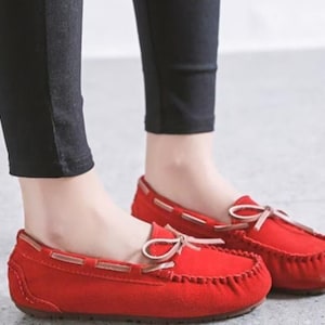 May include: Red suede moccasin-style flats with a rounded toe and a small bow detail. The shoes have a stitched sole and a leather-look trim. The flats are shown with black pants.