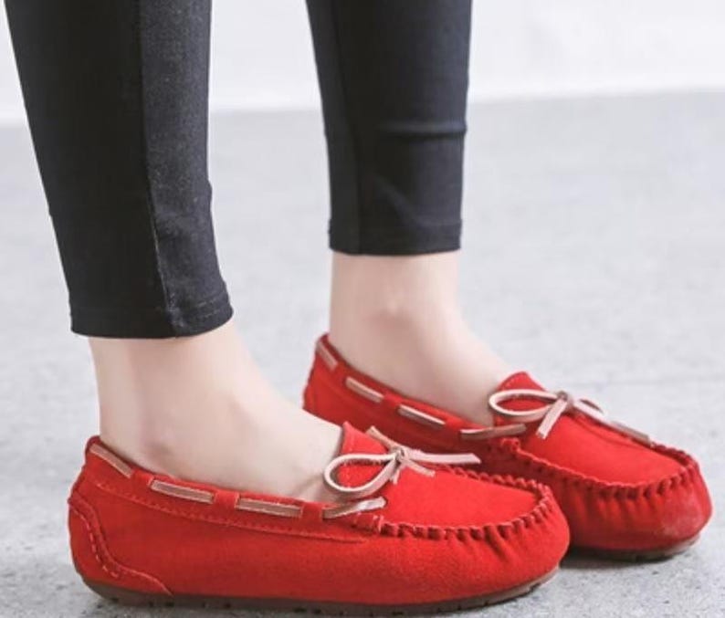 May include: Red suede moccasin-style flats with a rounded toe and a small bow detail. The shoes have a stitched sole and a leather-look trim. The flats are shown with black pants.