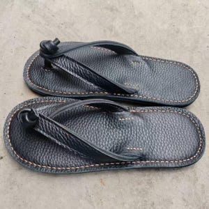 Puede incluir: Un par de chanclas de cuero negro. Las sandalias tienen una superficie texturizada y est&aacute;n cosidas con hilo marr&oacute;n claro alrededor de los bordes. Las correas est&aacute;n anudadas en la parte superior.