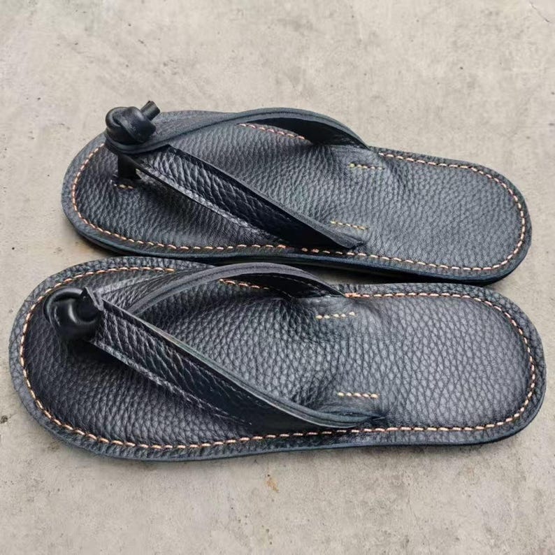 Puede incluir: Un par de chanclas de cuero negro. Las sandalias tienen una superficie texturizada y est&aacute;n cosidas con hilo marr&oacute;n claro alrededor de los bordes. Las correas est&aacute;n anudadas en la parte superior.