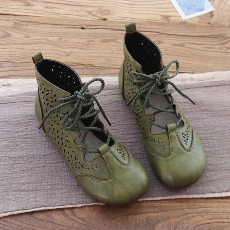 May include: A pair of olive green ankle boots with intricate cut-out patterns. The boots feature a lace-up design with matching green laces and a rounded toe. The shoes are made of a soft, textured material and appear to be leather.