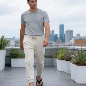 Puede incluir: Una persona con una camiseta gris de cuello redondo, pantalones deportivos color crema con cord&oacute;n y sandalias negras. La persona camina por un patio en la azotea con macetas blancas y un horizonte urbano al fondo.