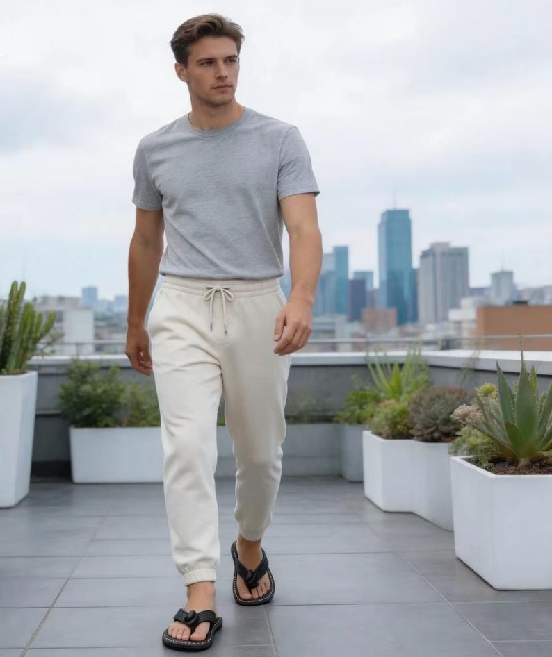 Puede incluir: Una persona con una camiseta gris de cuello redondo, pantalones deportivos color crema con cord&oacute;n y sandalias negras. La persona camina por un patio en la azotea con macetas blancas y un horizonte urbano al fondo.