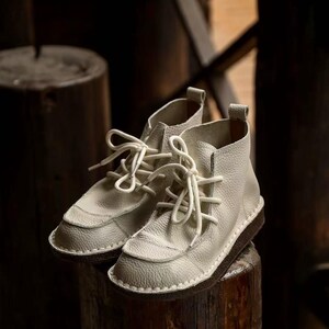 Pu&ograve; includere: Un paio di stivaletti alla caviglia in pelle bianco sporco con lacci bianchi. Gli stivaletti hanno una superficie strutturata e una suola marrone scuro. Le scarpe sono esposte su una superficie di legno, evidenziando il loro design.