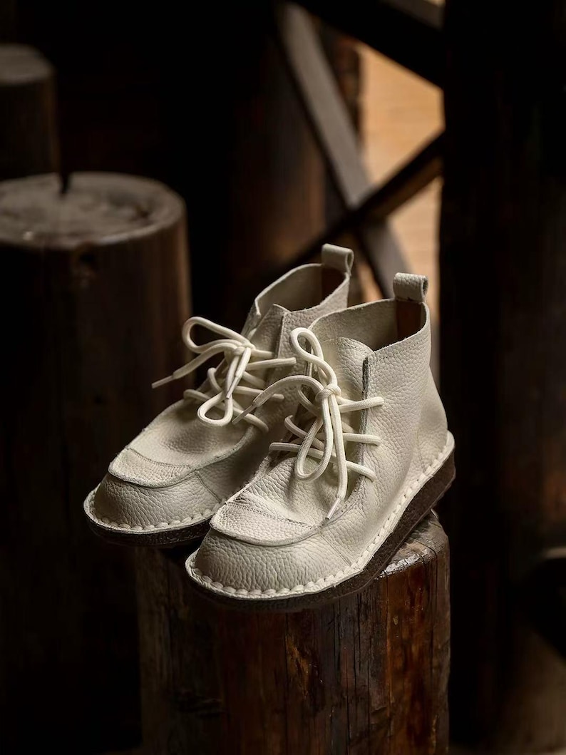 Pu&ograve; includere: Un paio di stivaletti alla caviglia in pelle bianco sporco con lacci bianchi. Gli stivaletti hanno una superficie strutturata e una suola marrone scuro. Le scarpe sono esposte su una superficie di legno, evidenziando il loro design.