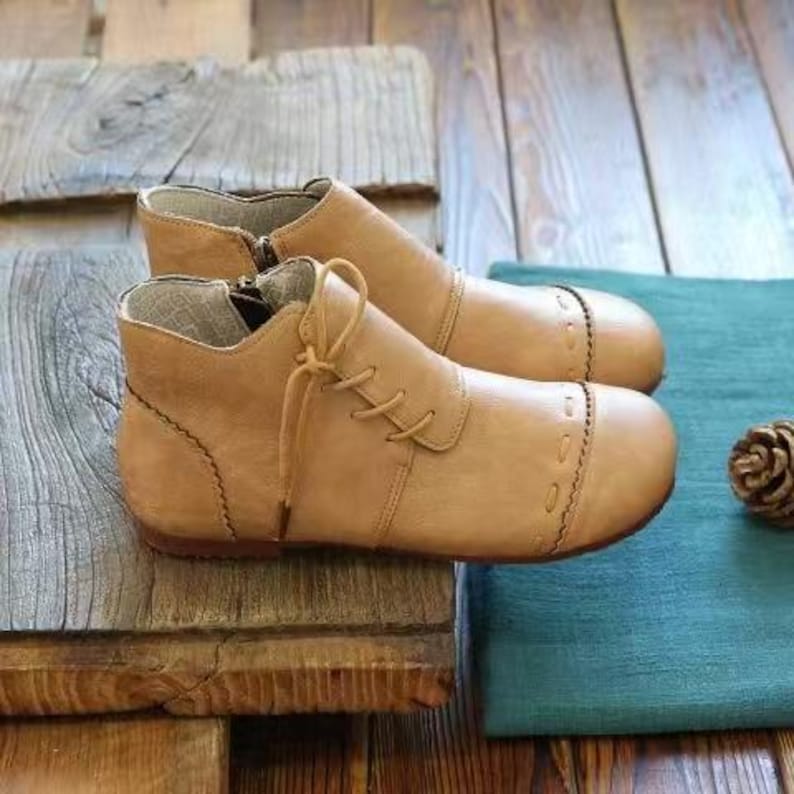 May include: A pair of tan leather ankle boots with laces and a zipper closure. The boots have decorative stitching around the toe and heel. They are placed on a wooden surface with a teal cloth and a pinecone in the background.