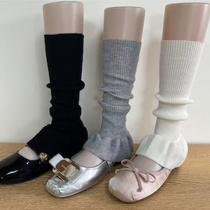 May include: Three pairs of leg warmers in black, gray, and white, displayed on mannequins with matching shoes. The black leg warmers are paired with black patent leather flats. The gray leg warmers are paired with silver flats. The white leg warmers are paired with pink flats.