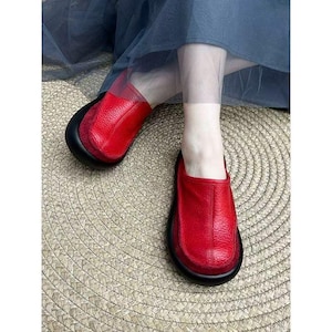 Puede incluir: Zapatos sin cordones de cuero rojo con suela negra. Los zapatos tienen una puntera redondeada y una parte superior de cuero texturizado. Los zapatos se muestran sobre una estera tejida. Los zapatos son de color rojo vibrante.