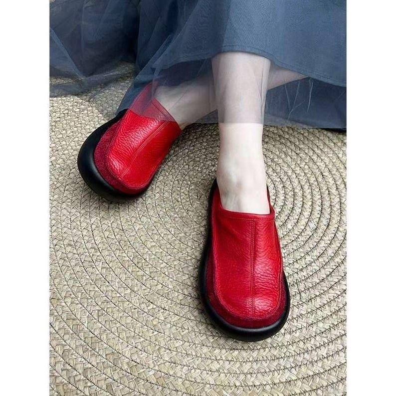 Puede incluir: Zapatos sin cordones de cuero rojo con suela negra. Los zapatos tienen una puntera redondeada y una parte superior de cuero texturizado. Los zapatos se muestran sobre una estera tejida. Los zapatos son de color rojo vibrante.