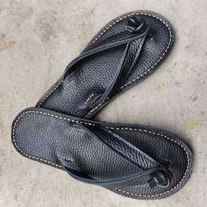Puede incluir: Un par de chanclas de cuero negro con una superficie texturizada. Las sandalias tienen un dise&ntilde;o de tanga con una correa entre los dedos. Los bordes est&aacute;n cosidos con hilo marr&oacute;n claro.