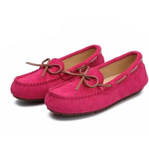 May include: A pair of bright pink suede moccasin-style shoes. Each shoe features a brown bow on the vamp and brown trim around the top. The shoes have a stitched design around the toe box and a flat sole.